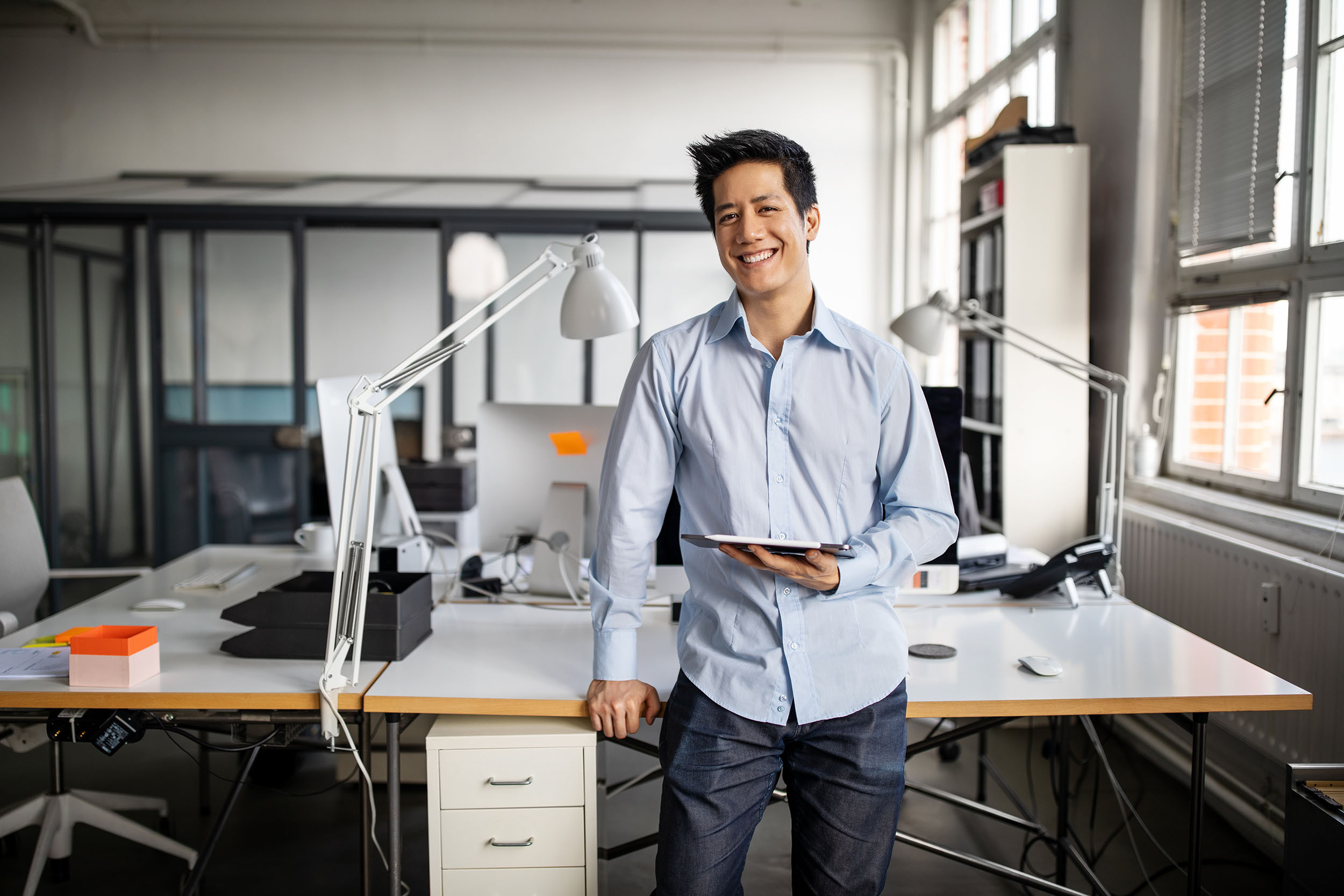 Male entrepreneur holding a tablet grins while sitting on his home office desk.