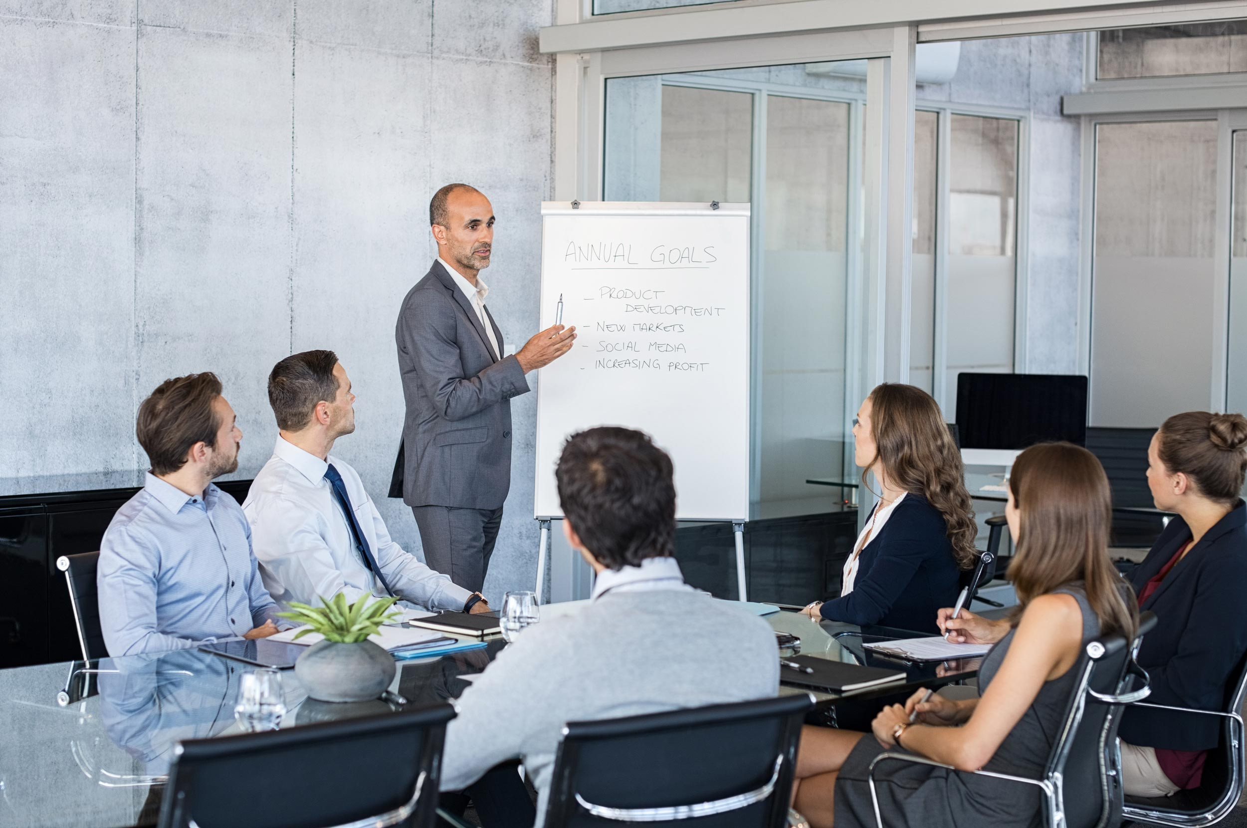 One coworker goes over the agenda on a whiteboard with his other seated coworkers in a business meeting
