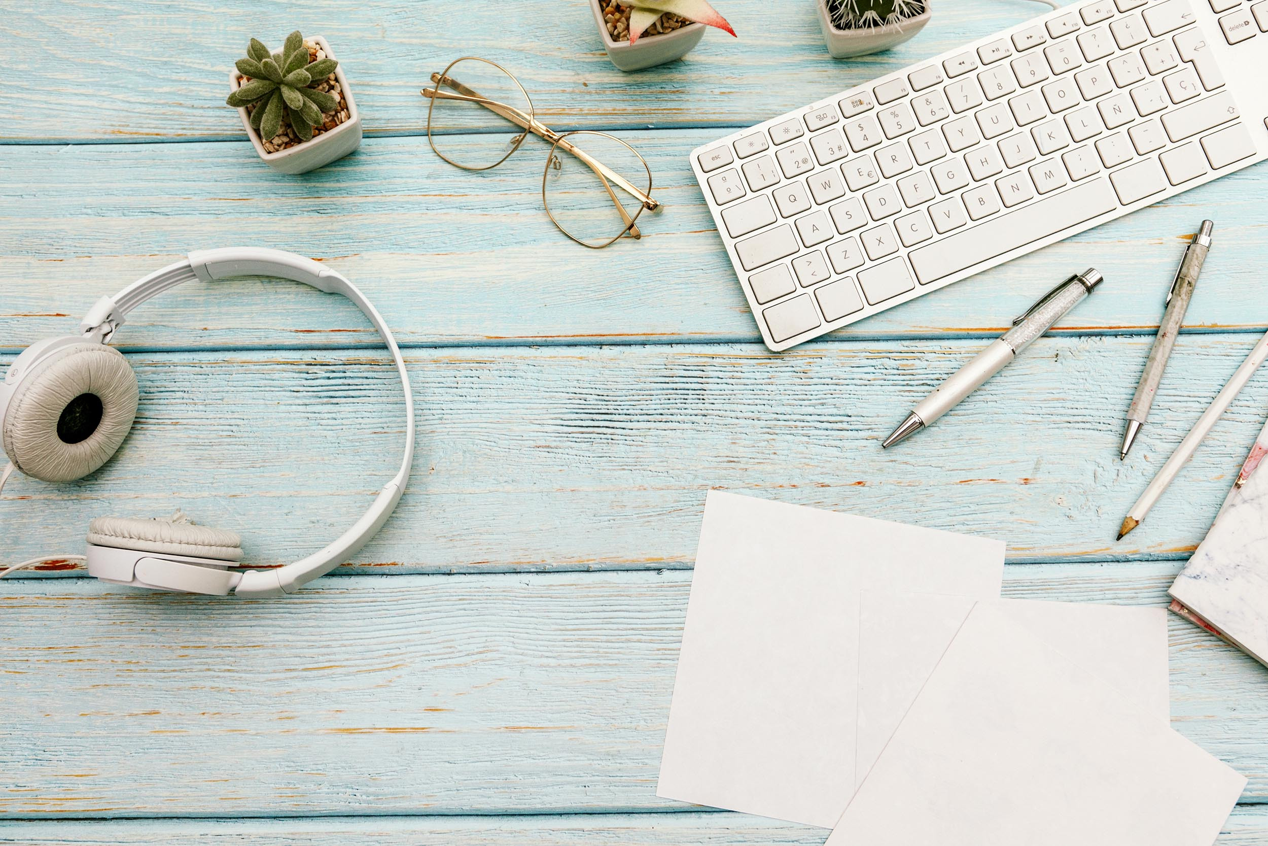 flatlay of computer headphone eyeglass pen plant and papers on the wood