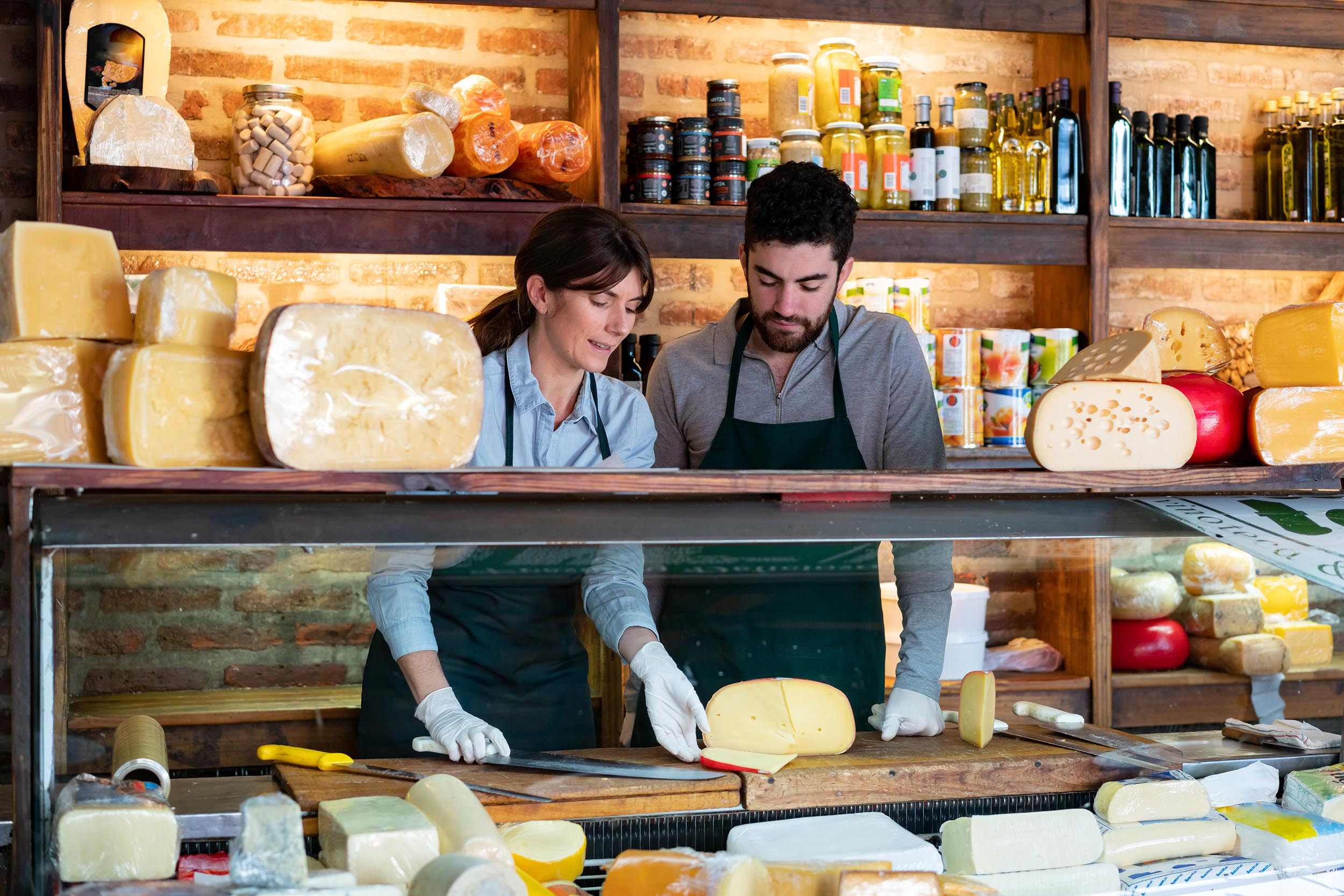 a deli shop owner teaching employee how to cut cheese